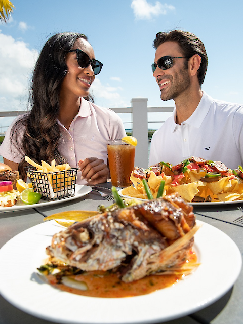 A couple is enjoying a sunny outdoor meal with dishes including nachos, fries, and fish, accompanied by a drink, all while smiling.