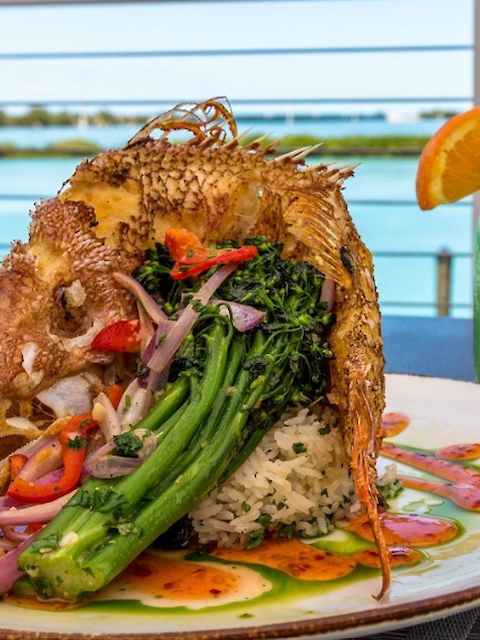 A whole cooked fish with vegetables and rice is served on a plate, paired with a green drink garnished with an orange slice.