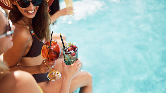 Two people sit poolside in swimsuits, holding colorful drinks with fruit garnishes. The sun is shining, creating a relaxed summer atmosphere.