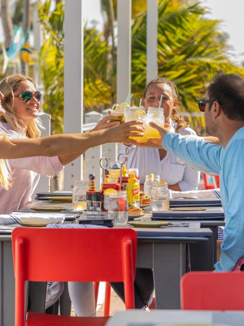 A group of people clinks drinks at an outdoor restaurant table, surrounded by palm trees, with a vibrant and sunny atmosphere.