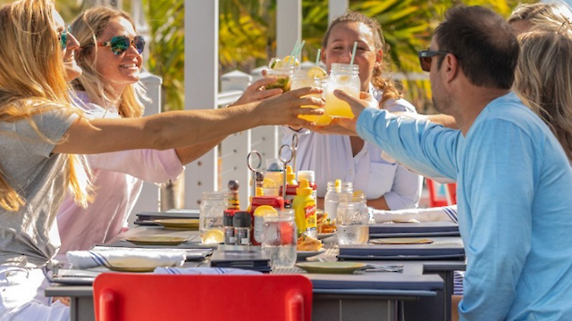 A group of people clinks drinks at an outdoor restaurant table, surrounded by palm trees, with a vibrant and sunny atmosphere.