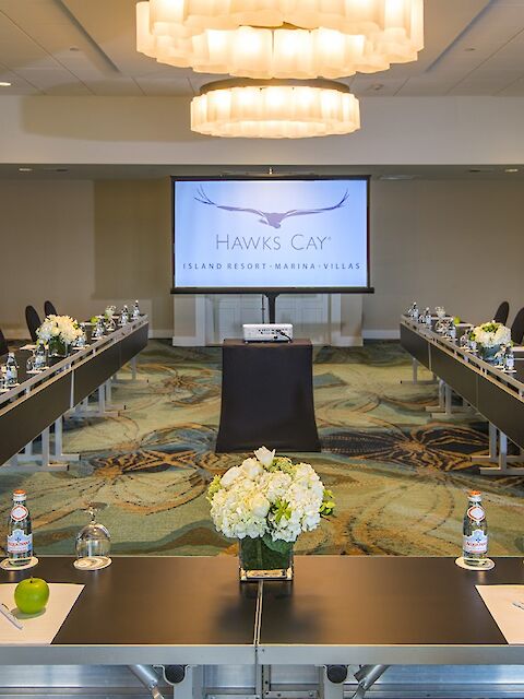 This image shows a conference room set up with U-shaped tables, chairs, water bottles, apples, and flowers, facing a screen displaying "Hawks Cay."