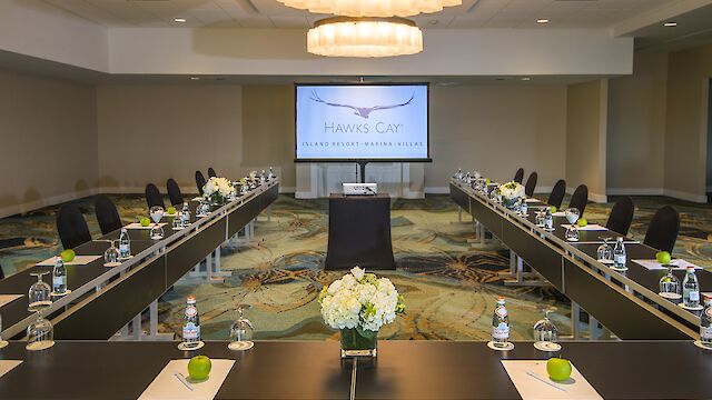 This image shows a conference room set up with U-shaped tables, chairs, water bottles, apples, and flowers, facing a screen displaying "Hawks Cay."