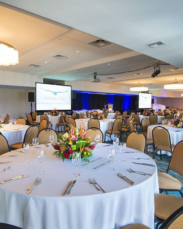 The image shows a decorated event hall with round banquet tables, floral centerpieces, chairs, and presentation screens.