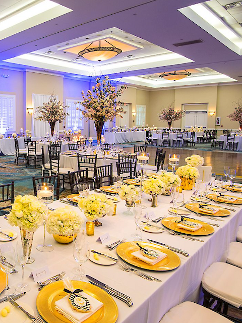 Elegant banquet hall with long tables, yellow-themed decor, floral centerpieces, and candles. Tables set for a formal event with plates and glasses.