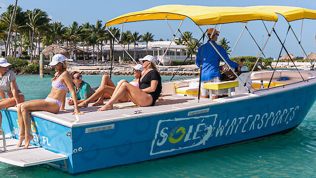 A boat labeled "Sole Watersports" with four people in swimwear relaxing on it in turquoise water, with palm trees and a beach in the background.