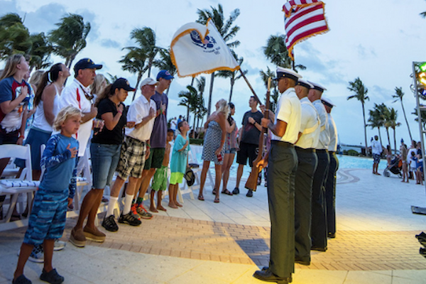 A military ceremony with people standing and respectful, soldiers holding flags, and palm trees in the background near a poolside location.