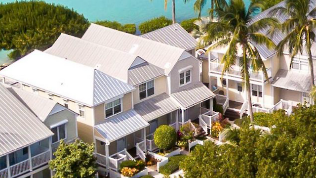 The image shows a tropical residential area with white-roofed houses, surrounded by lush greenery and palm trees near a body of water, ending the sentence.