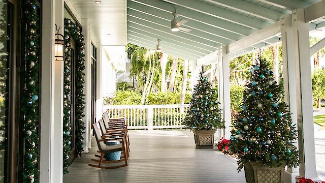 A decorated porch with Christmas trees, rocking chairs, wrapped columns, and festive greenery, with a view of lush greenery outside the porch.