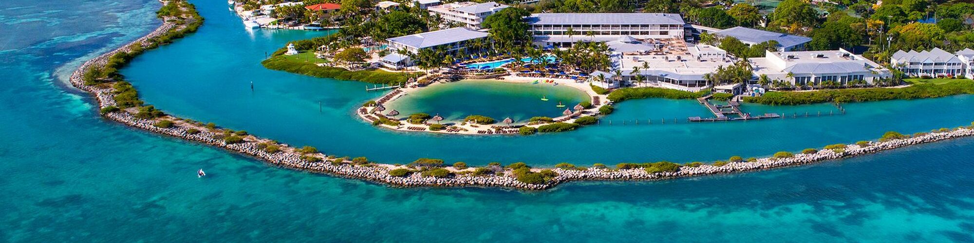 Aerial view of a coastal resort island surrounded by turquoise blue waters, with buildings, greenery, and a central sandy beach area.