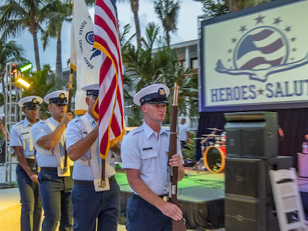 A group of uniformed individuals holding flags and a rifle march beside a stage displaying a "Heroes Salute" sign, with palm trees in the background.