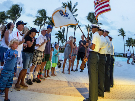 A group of people stands while uniformed personnel hold flags near a pool surrounded by palm trees during a ceremony or event.