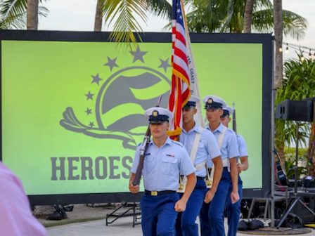 Four uniformed individuals are carrying a flag, marching in front of a large screen displaying the word "HEROES" and a logo, with palm trees in the background.