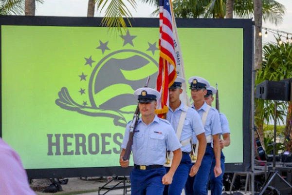 Four uniformed individuals are carrying a flag, marching in front of a large screen displaying the word "HEROES" and a logo, with palm trees in the background.