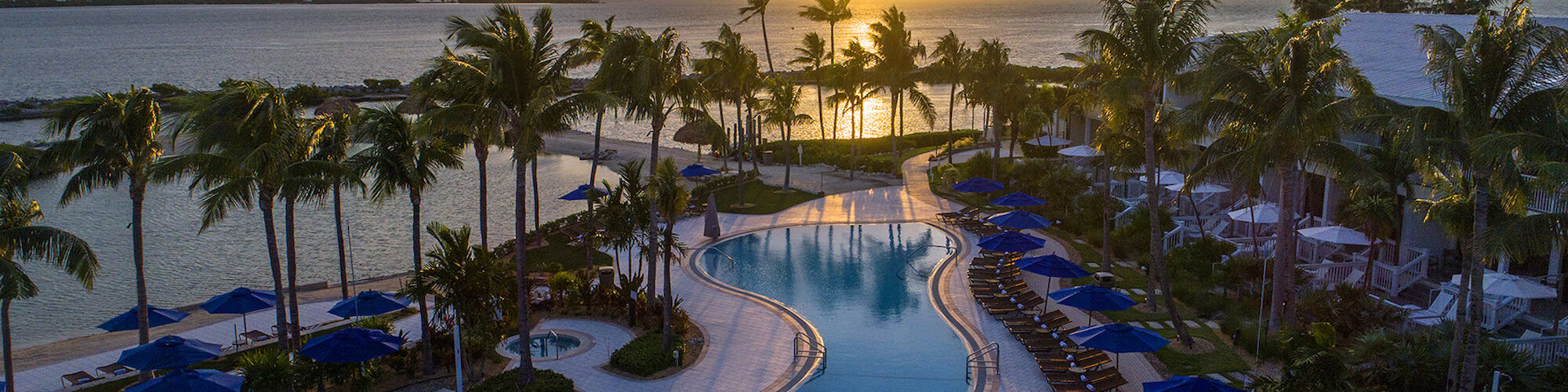 A resort pool is surrounded by palm trees and lounge chairs, with a sunset over the ocean in the background, creating a tropical scene.