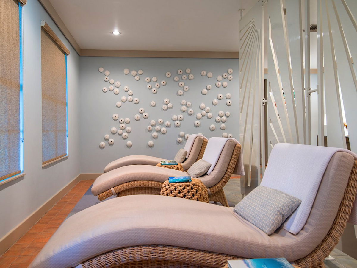 The image shows a relaxing spa room with wicker lounge chairs, pillows, and a wall decorated with small white spherical elements.