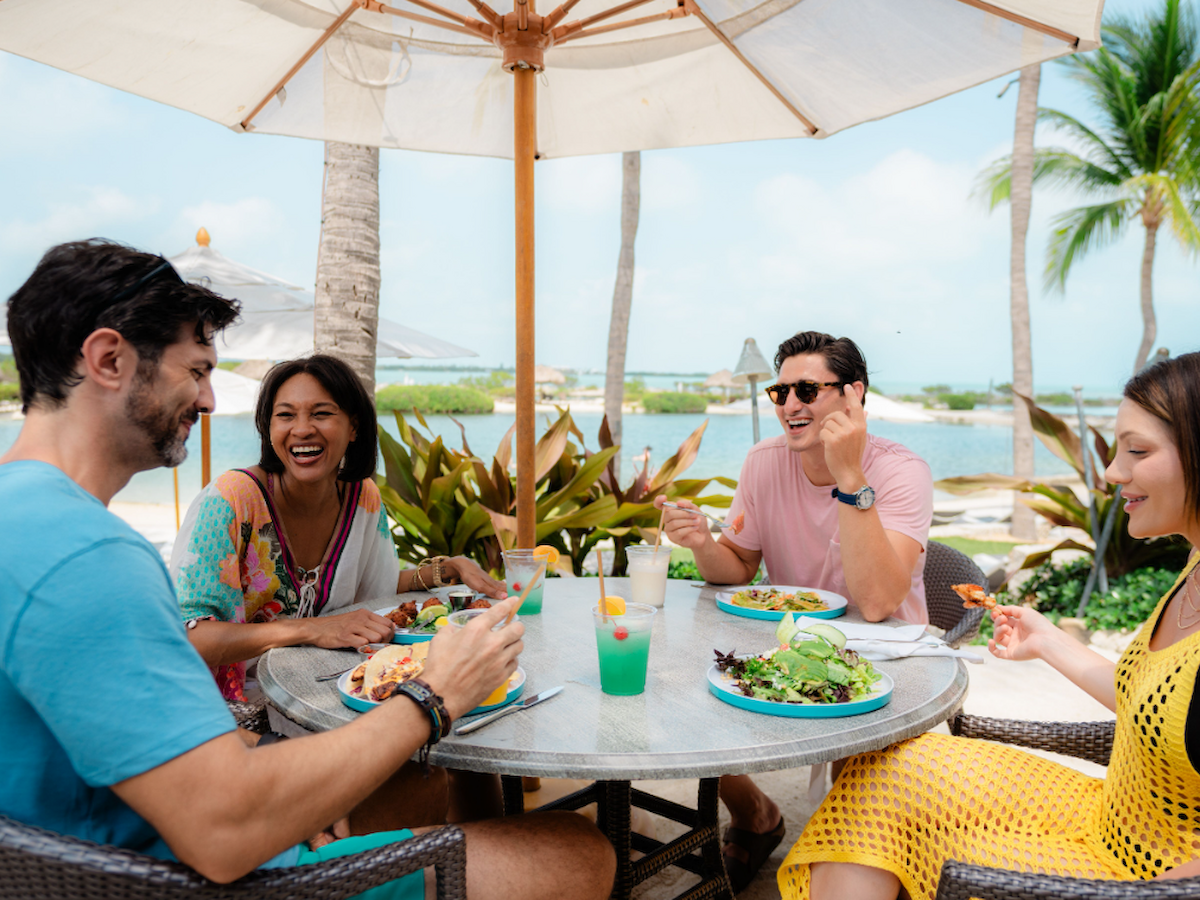 A group of four people enjoying a meal at an outdoor table by the beach, with palm trees and a scenic waterfront view in the background.