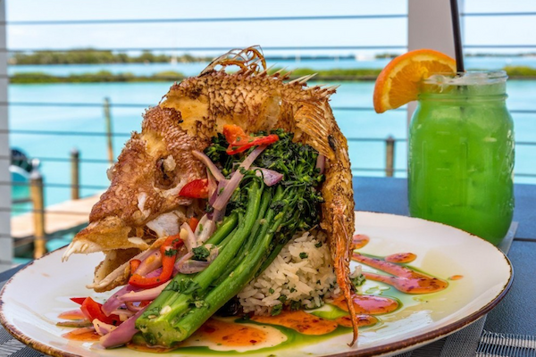 A plated whole fish garnished with vegetables sits beside a green cocktail, overlooking a water view.