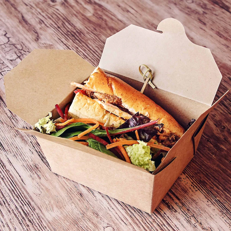 The image shows a sandwich with mixed greens and sliced vegetables in an open cardboard takeout box on a wooden surface.