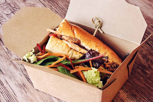 A sandwich with mixed greens and vegetables in a rectangular cardboard takeout box on a wooden surface.