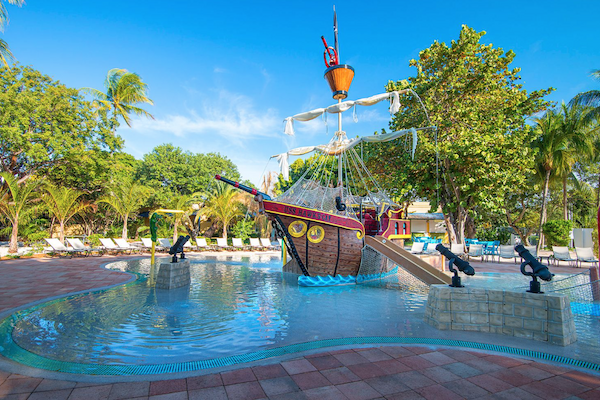 A children's water play area with a pirate ship theme, surrounded by trees and lounge chairs under a clear blue sky.