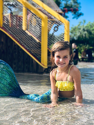 A child in a yellow swimsuit with a mermaid tail smiles while sitting in shallow water at a play area with nets and slides nearby.