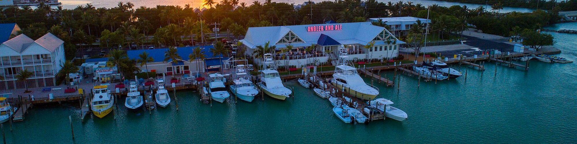 Aerial view of a marina at sunset with boats docked, calm water, palm trees, and buildings along the shoreline under an orange sky.