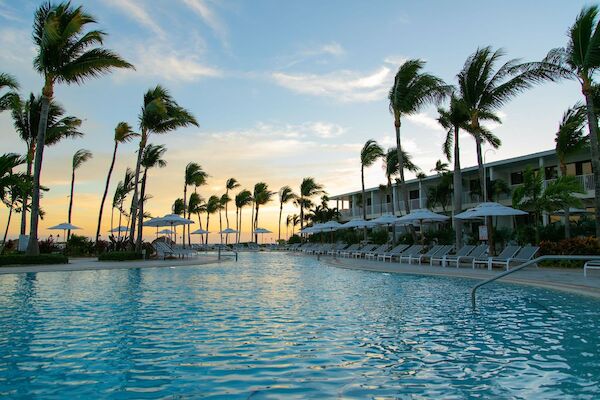 A serene poolside scene with palm trees, lounge chairs, umbrellas, and a building in the background during sunset.