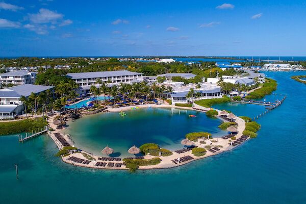 Aerial view of a tropical resort with buildings, a lagoon, beaches, and lush greenery surrounded by clear blue water under a sunny sky.