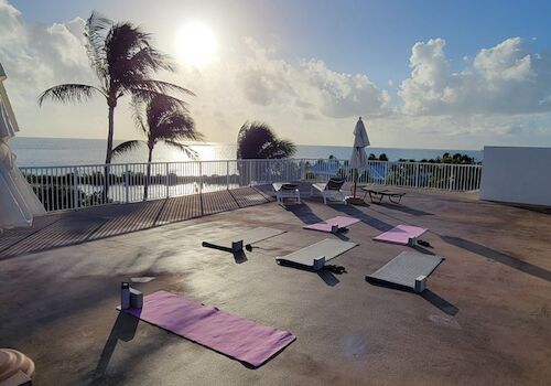 Rooftop setup with yoga mats facing the ocean under a clear sky, palm trees in the breeze, and the sun shining brightly.