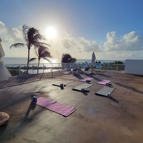 Rooftop setup with yoga mats facing the ocean under a clear sky, palm trees in the breeze, and the sun shining brightly.