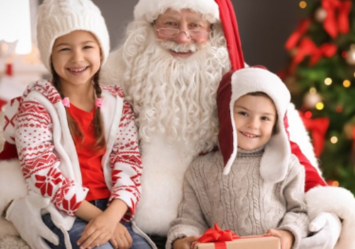 A person dressed as Santa Claus is sitting with two smiling children in festive outfits, near a decorated Christmas tree, holding a gift.