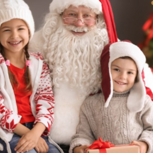 A person dressed as Santa Claus is sitting with two smiling children in festive outfits, near a decorated Christmas tree, holding a gift.