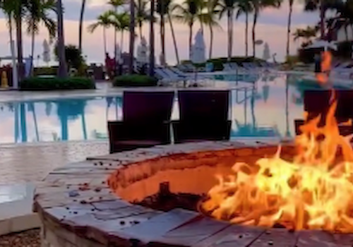 A fire pit is lit up near a swimming pool surrounded by palm trees and lounge chairs, creating a cozy and tropical atmosphere.