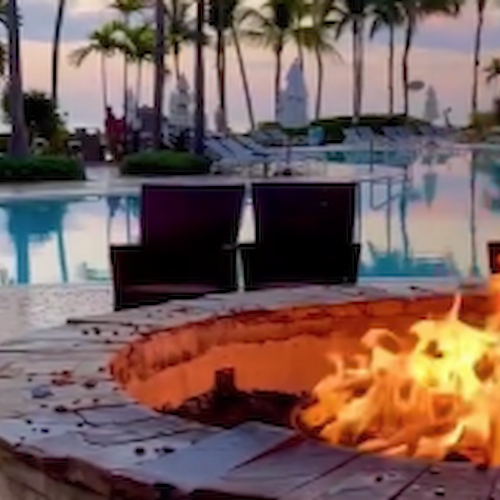 A fire pit is lit up near a swimming pool surrounded by palm trees and lounge chairs, creating a cozy and tropical atmosphere.