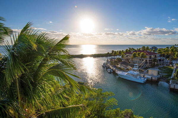 A scenic view of a waterfront, with a tranquil sea, boats docked, palm trees, and a vibrant sunset.