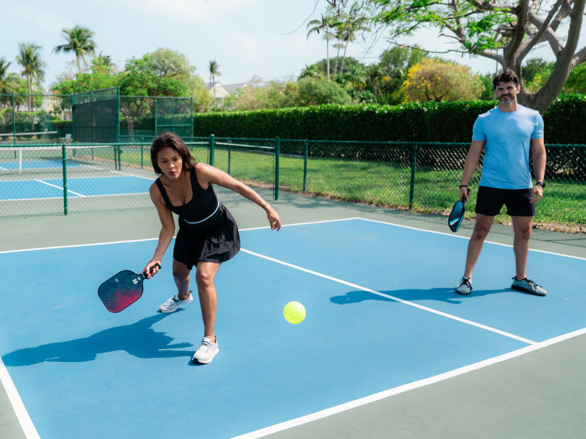 Two people are playing pickleball on an outdoor court. One player is about to hit the ball with a paddle.