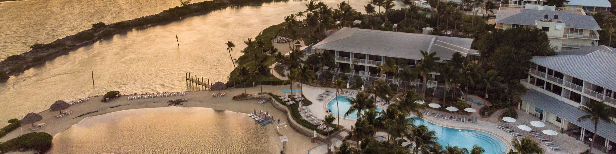The image shows a beachfront resort at sunset with pools, palm trees, and surrounding ocean views.
