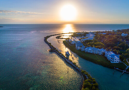 Aerial view of a coastal area at sunset, with buildings, trees, and a winding waterway leading to the ocean.