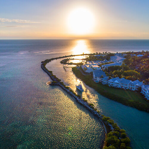 Aerial view of a coastal area at sunset, with buildings, trees, and a winding waterway leading to the ocean.