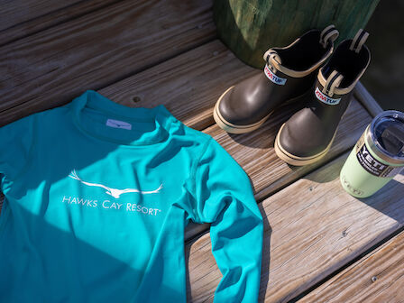 A turquoise Hawks Cay Resort shirt, a pair of waterproof boots, and a Yeti tumbler are placed on wooden decking.