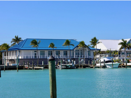 A blue-roofed building with palm trees sits by the waterfront, with boats docked nearby and clear blue skies above.