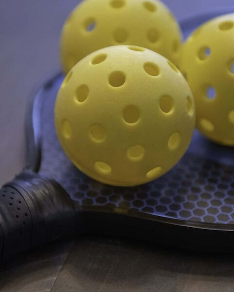 The image shows a pickleball paddle and three yellow perforated balls placed on top of it.