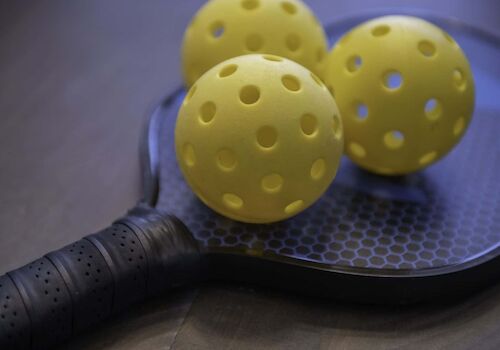 The image shows a pickleball paddle and three yellow perforated balls placed on top of it.