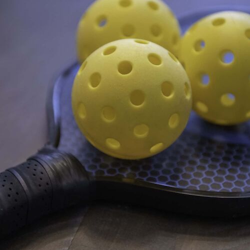 The image shows a pickleball paddle and three yellow perforated balls placed on top of it.