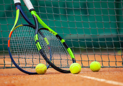 Two tennis rackets are leaning against a net with four tennis balls on a clay court.