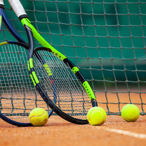 Two tennis rackets are leaning against a net with four tennis balls on a clay court.