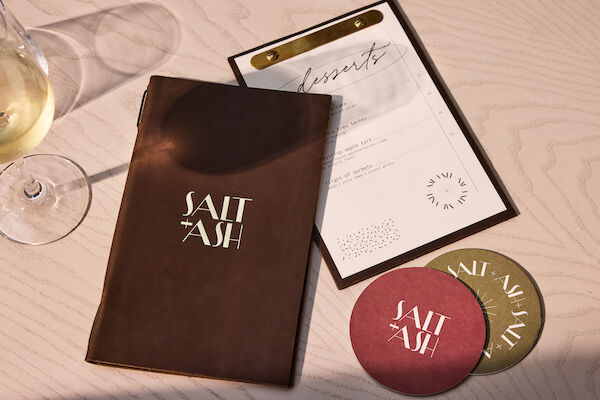 A wine glass, a leather-bound menu labeled "Salt + Ash," a dessert list, and two coasters sit on a light wood table.