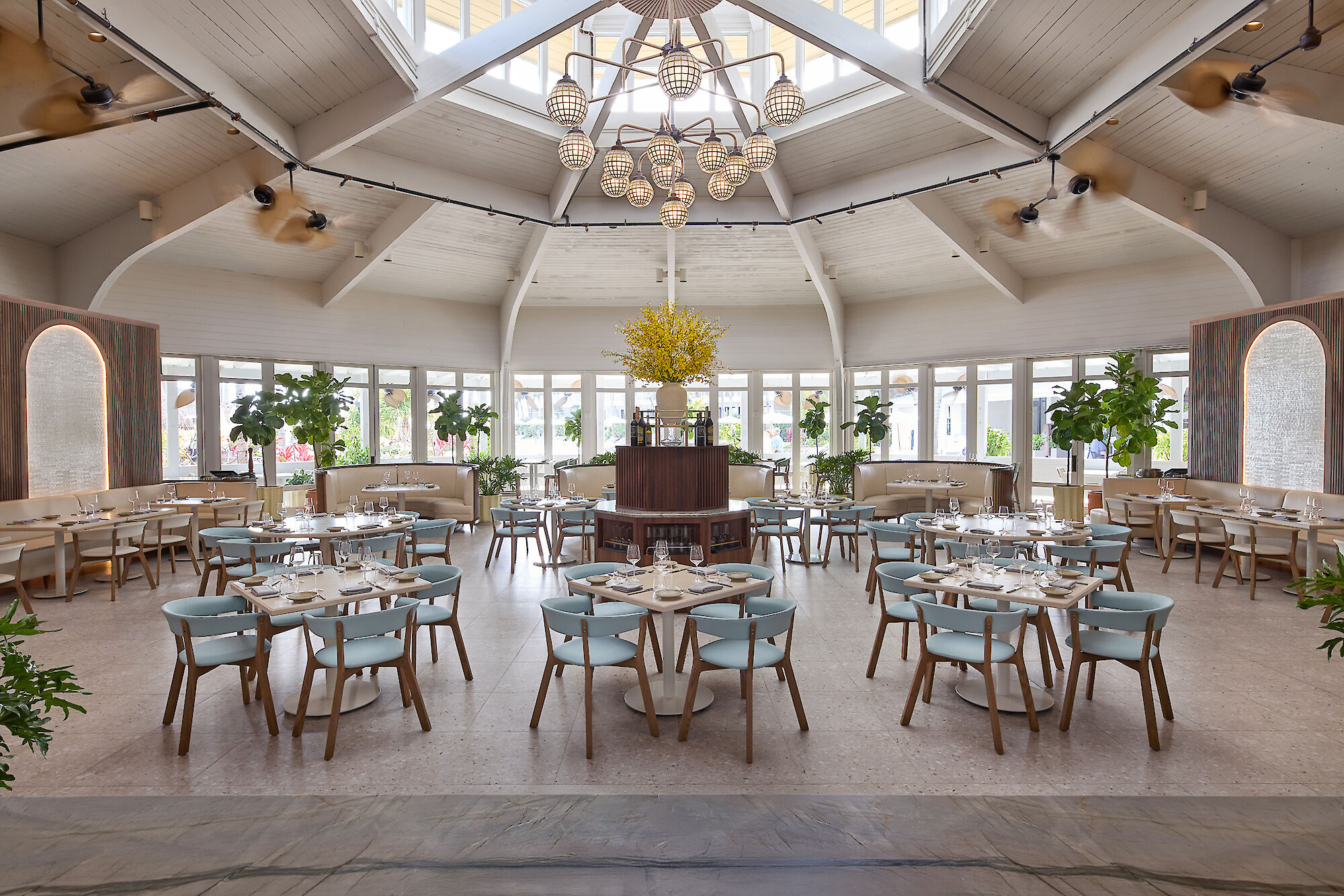 A spacious, elegant restaurant interior with light blue chairs, round tables, yellow floral centerpiece, large windows, and modern lighting.