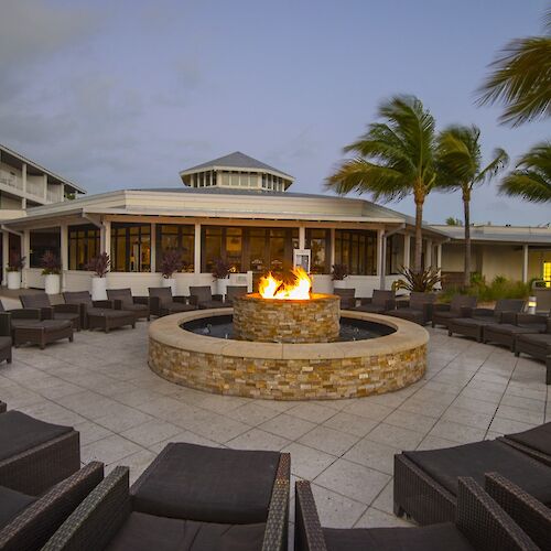 An outdoor area with a central fire pit surrounded by chairs, with palm trees and a building in the background under a clear sky.